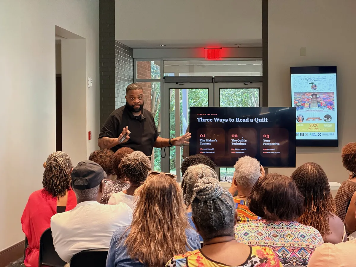 Wesley Hall speaking from a podium at the 2026 Atlanta Quilt Festival with a quilt image projected on the screen behind him