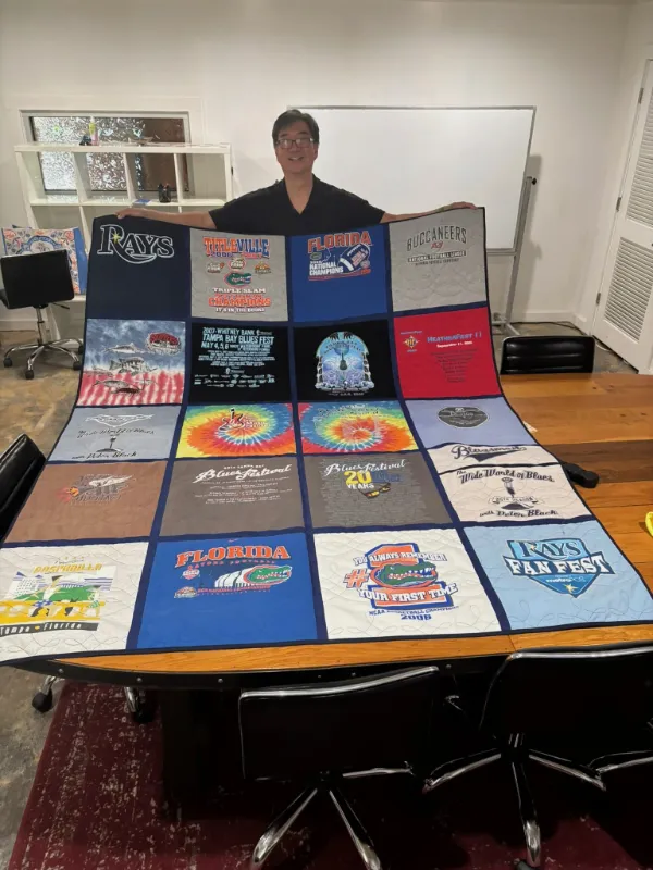 Client displaying a large custom Florida sports and concert t-shirt quilt featuring Tampa Bay Rays, Buccaneers, and Gators shirts, handmade in Atlanta by Quilts by Big Wes