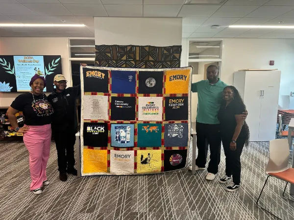 Four Emory University alumni and students holding a custom Emory t-shirt quilt with kente cloth sashing, made by Quilts by Big Wes in Atlanta