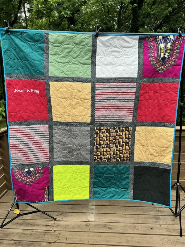 Memory quilt made from colorful clothing including dashiki fabric, faith shirts, and bold solid colors in teal, red, gold, and black, displayed outdoors