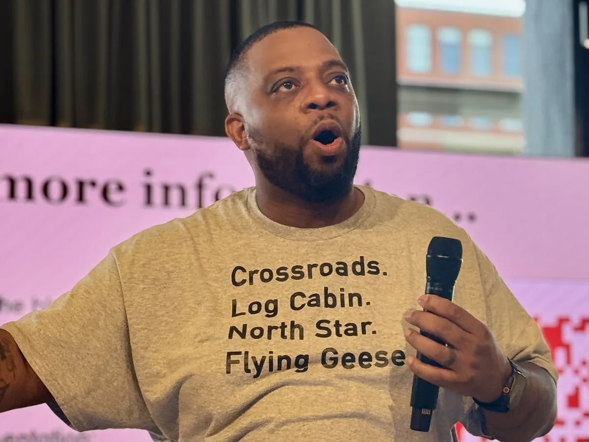 Wesley Hall speaking at a quilt conference in a t-shirt listing traditional quilt block names: Crossroads, Log Cabin, North Star, Flying Geese